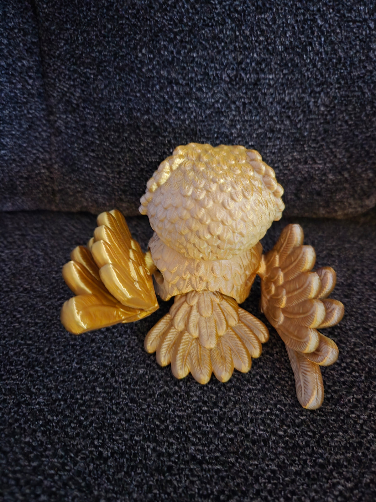A small sculptural figurine of a bird, likely a goldfinch, with textured cream-colored body and wings and bright gold-colored wings, displayed on a dark textured fabric background.