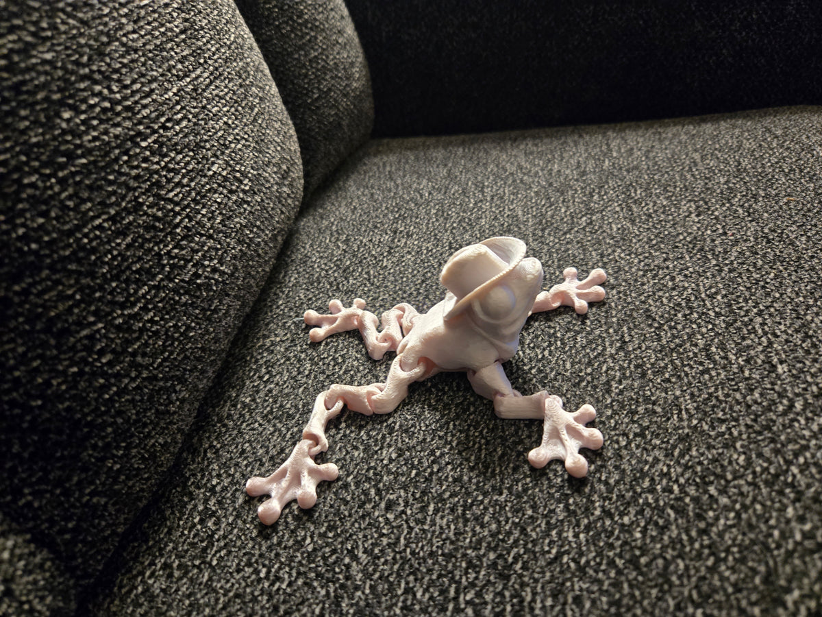 A small white frog figurine wearing a small hat, posed on a dark gray textured fabric surface next to a dark gray upholstered chair.