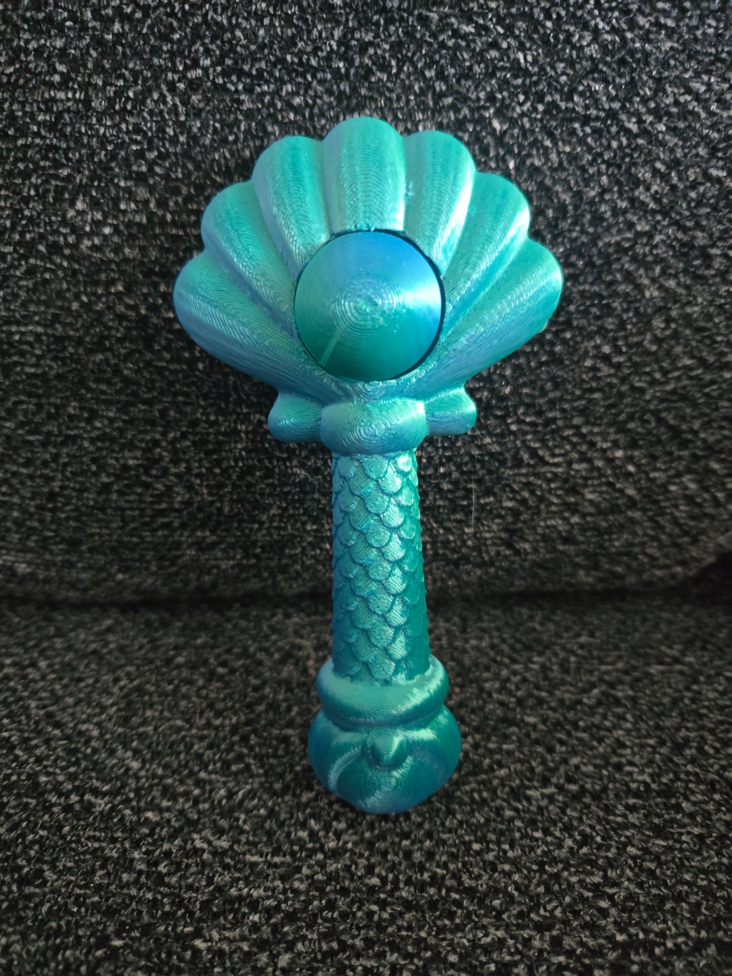 A turquoise and teal mermaid-themed rattle with a scalloped shell top, a round bead, and a textured stem resembling a mermaid tail, resting on a dark gray textured fabric background.