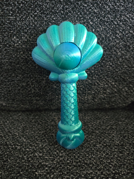 A turquoise and teal mermaid-themed rattle with a scalloped shell top, a round bead, and a textured stem resembling a mermaid tail, resting on a dark gray textured fabric background.